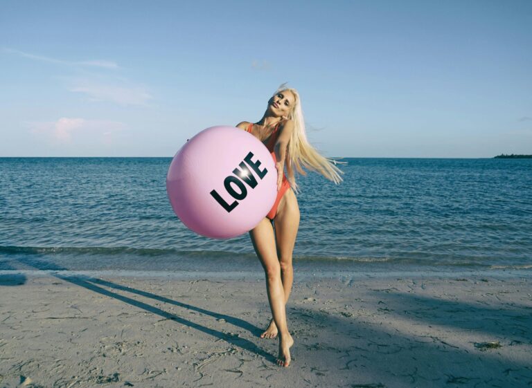 Eine blonde Frau im Bikini hält einen großen, rosa Ball vor ihrem Körper auf dem "Love" steht.