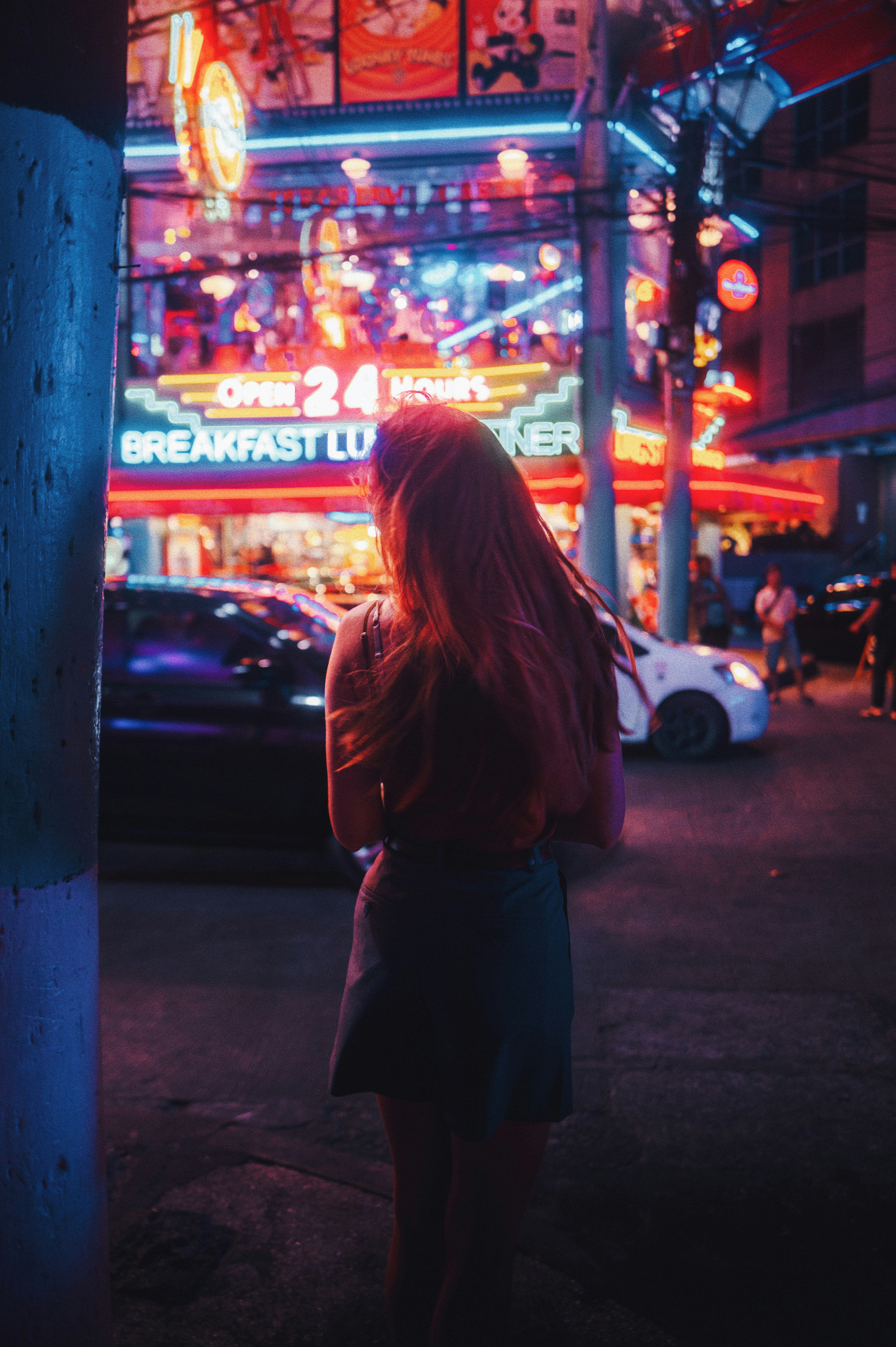Blick von hinten auf eine Frau, die nachts vor einem mit Neon-Lichtern beleuchteten Gebäude steht.