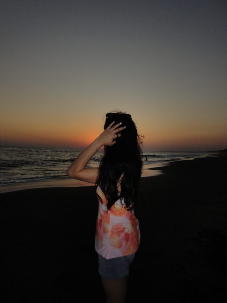 Eine junge Frau in einem bunten Kleid vor einem Sonnenuntergang am Strand.